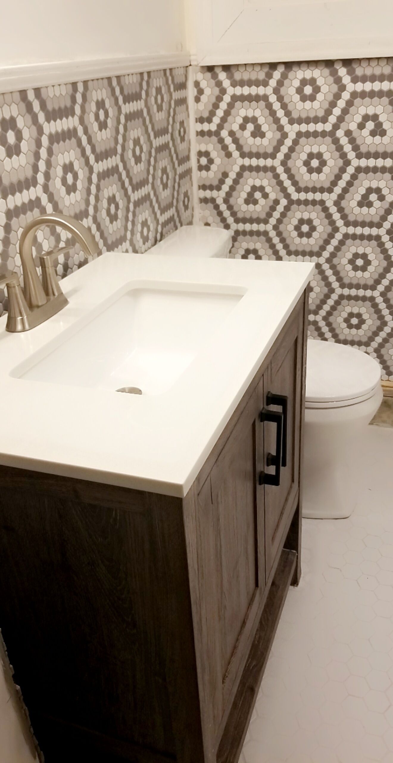 A bathroom with a white sink, dark wooden cabinet, and a toilet. The walls have a hexagonal patterned tile design.