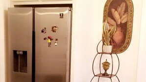 A kitchen with a stainless steel refrigerator, a small metal shelf with a vase, and two wooden stools.