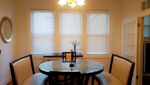 A dining area with a round glass table, four chairs, and three windows with blinds.