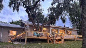 A two-story house with a deck, outdoor lighting, and a large tree. - Cozy Cottage on Crooked Lake (Babson Park)
