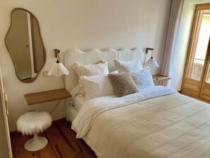 A bedroom with a bed, bedside tables, a mirror, a fluffy stool, and a window with curtains.
