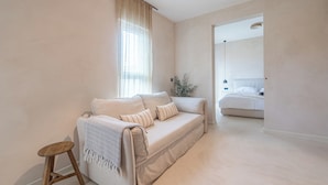 A neutral-toned living room with a sofa, a small wooden stool, and a window with sheer curtains.