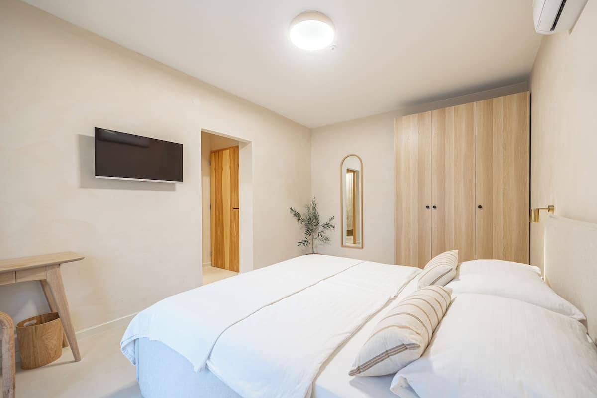 A bedroom with a bed, a TV mounted on the wall, a wooden wardrobe, and a small desk with a chair.
