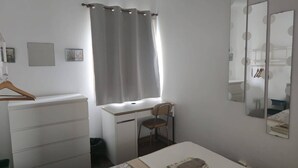 Een slaapkamer met een bed, een bureau, een stoel, een kledingkast, een spiegel en een raam met gordijnen.
