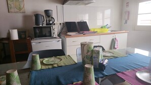 Een keuken met een magnetron, koffiezetapparaat en een kleine koelkast. Er staat een eettafel met een blauwe tafelkleed en verschillende keukenbenodigdheden erop.