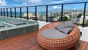 Een ronde rieten bank met kussens, een houten terras en een stadszicht door grote ramen.