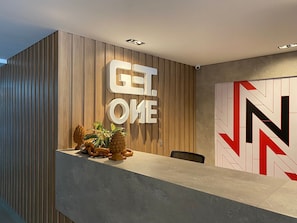 A reception desk with a wooden panel wall, a large 'GET ONE' sign, and a geometric wall art piece. - Bela Hospedagem - Get One Bessa (João Pessoa)