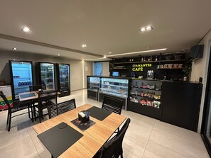 A café with a counter displaying snacks and beverages, a seating area with tables and chairs, and vending machines.