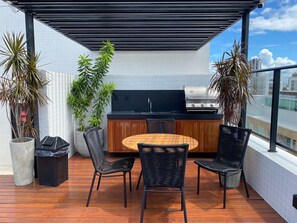 A rooftop patio with a round wooden table, four black wicker chairs, a grill, and potted plants. - Bela Hospedagem - Get One Bessa (João Pessoa)