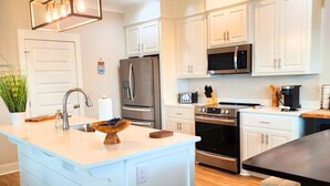 A modern kitchen with white cabinetry, stainless steel appliances, and a central island with a sink and bowl of fruit. - Boating, Fishing, Kayaking and more at this NEW Waterfront Bay St. Louis Retreat (Bay St. Louis)