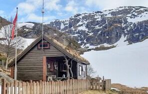 Uma cabana de madeira com telhado de palha, cercada por uma vedação de madeira, com montanhas nevadas ao fundo.