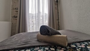 Una cama con una toalla enrollada y una manta, una ventana con cortinas y una lámpara.