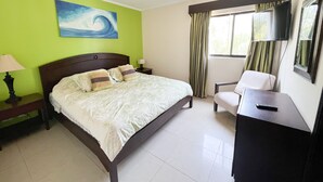 A hotel room with a bed, a desk, a chair, a ceiling fan, a painting on the wall, and a window with curtains. - 2BR/2BR w/Rooftop Terrace, steps from the Beach, Tamarindo Condo (Tamarindo)