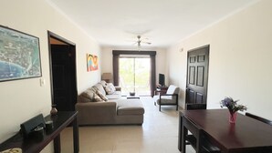 A living room with a sofa, a dining table, and a TV. There is a ceiling fan and a window with curtains. - 2BR/2BR w/Rooftop Terrace, steps from the Beach, Tamarindo Condo (Tamarindo)