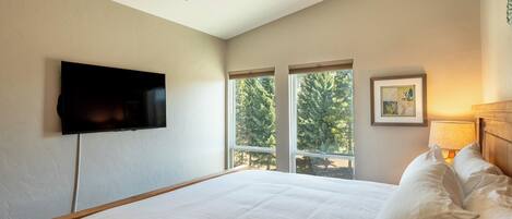 A bedroom with a bed, a television mounted on the wall, and two windows with a view of trees.