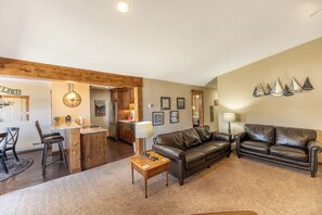 A living room with a brown leather couch, a wooden coffee table, and a kitchen area visible through an open doorway. - Lake Cliffe #303, Building E By Summit County Mountain Retreats (Dillon)