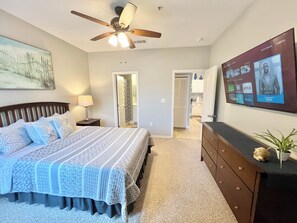 A bedroom with a bed, a dresser, a ceiling fan, a TV mounted on the wall, and a door leading to another room. - 2-104 Legacy Dunes Resort, Ground Floor Lake View, Heated Pool,Gym (Kissimmee)
