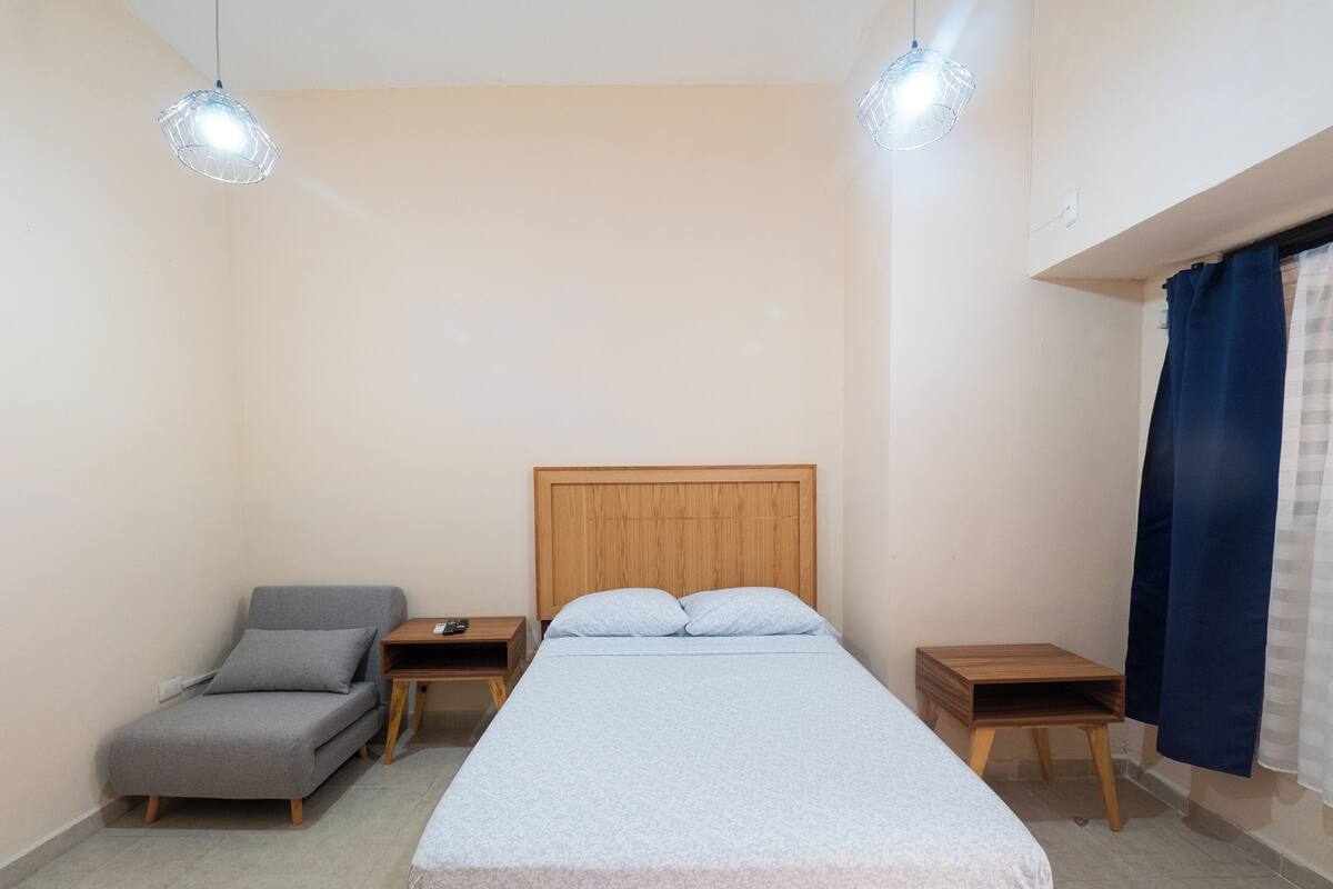 A bedroom with a bed, a chair, two bedside tables, and a hanging light fixture.