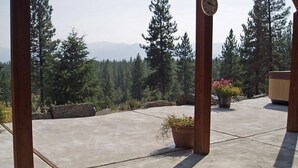 A concrete patio with wooden posts, a potted plant with pink flowers, and a view of a forested mountain range. - Rocky Mountain Getaway (Plains)