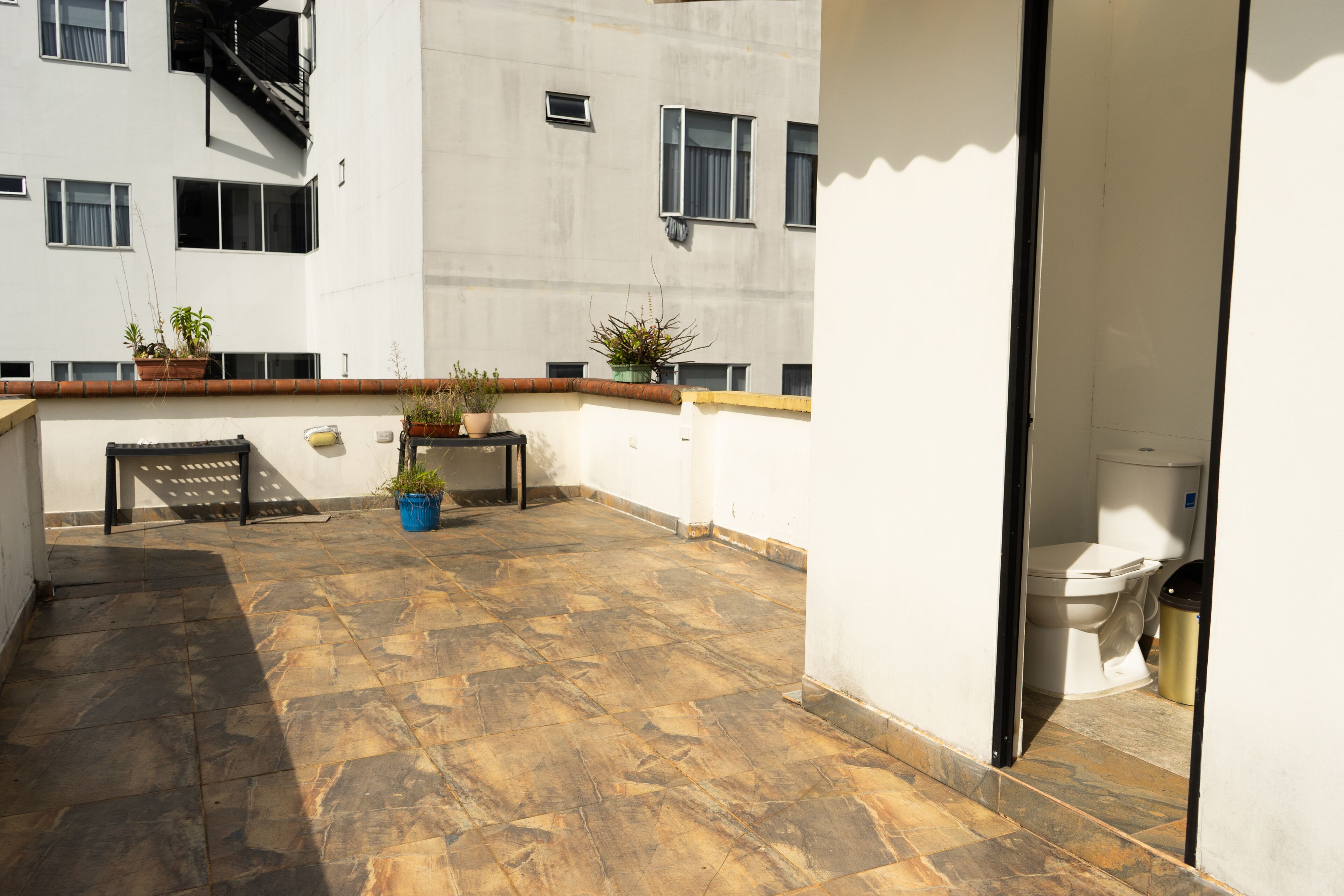 A rooftop terrace with stone flooring, potted plants, a bench, and a view of a building with multiple windows.