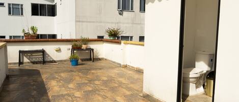 A rooftop terrace with stone flooring, potted plants, a bench, and a view of a building with multiple windows.