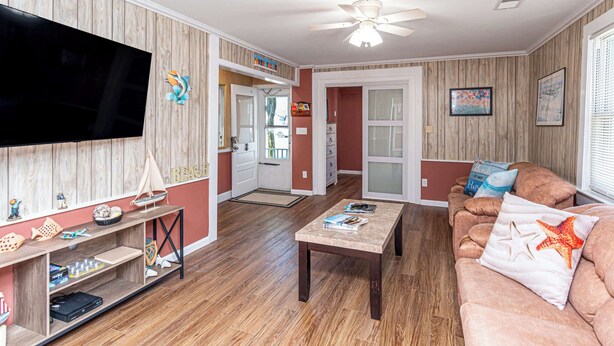 A living room with a flat-screen TV, wooden flooring, a ceiling fan, a sofa with beach-themed pillows, and a coffee table.