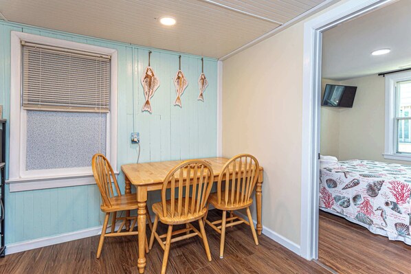 A dining area with a wooden table and chairs, a window with blinds, and a bed with a patterned bedspread.