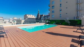 A rooftop pool with wooden decking and lounge chairs, surrounded by a cityscape and a building with a green wall and air conditioning units. - Paseo de las Cisternas (Buenos Aires)