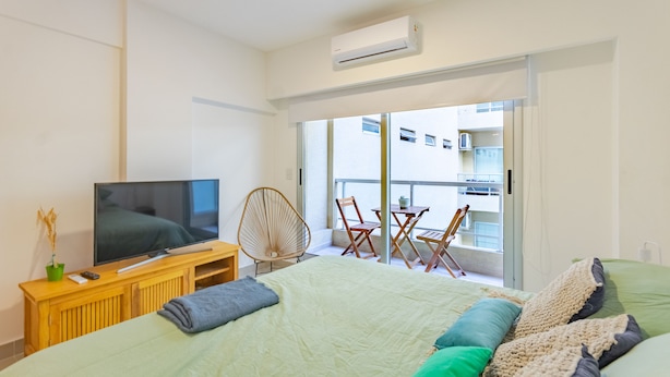 A bedroom with a bed, a TV on a wooden stand, a chair, a table, and a balcony with a view of buildings. - Paseo de las Cisternas (Buenos Aires)