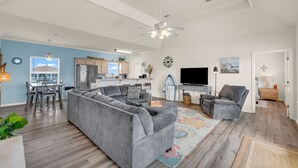 A spacious living room with a gray sectional sofa, a gray armchair, and a flat-screen TV. A ceiling fan and a dining area with a table and chairs are visible. - Beach Blessing with ocean view, 3br/2bath sleeps 8 (Bolivar Peninsula)