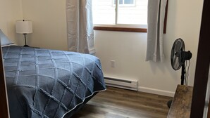 Una camera da letto con un letto, un comodino, una lampada, una finestra con tende, una ventola da terra e un tavolo di legno.