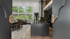 Une cuisine moderne dotée d’un îlot central, de grandes fenêtres et d’un coin repas avec des chaises en bois et un tapis.