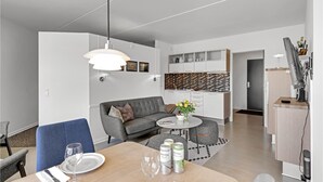 Un salon moderne avec un canapé gris, une table à manger avec deux verres à vin et des canettes, une table basse ronde, et une cuisine en arrière-plan.