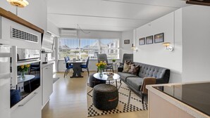 Une cuisine moderne avec des meubles blancs, un coin repas avec des chaises bleues, un canapé gris et une grande fenêtre donnant sur des immeubles.