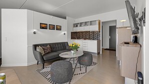Un salon moderne avec un canapé gris, une table basse ronde et des poufs gris. On y trouve également un coin cuisine avec des meubles blancs et un pare-feu noir et blanc.