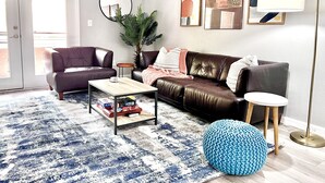 Un salon avec un canapé en cuir marron, une table basse, un pouf bleu en tricot et une lampe sur pied.