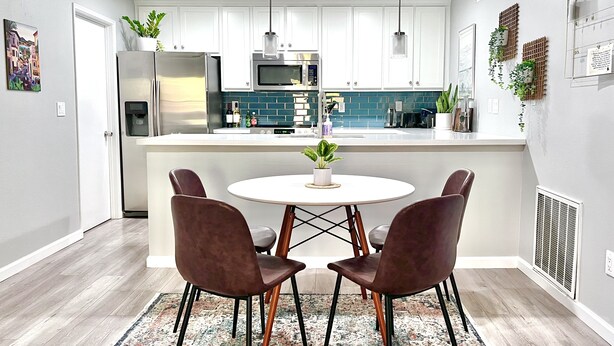 Une cuisine moderne avec des armoires blanches, un réfrigérateur en acier inoxydable, un four à micro-ondes et un évier. On y trouve une table ronde avec quatre chaises, et une petite plante en pot posée dessus.