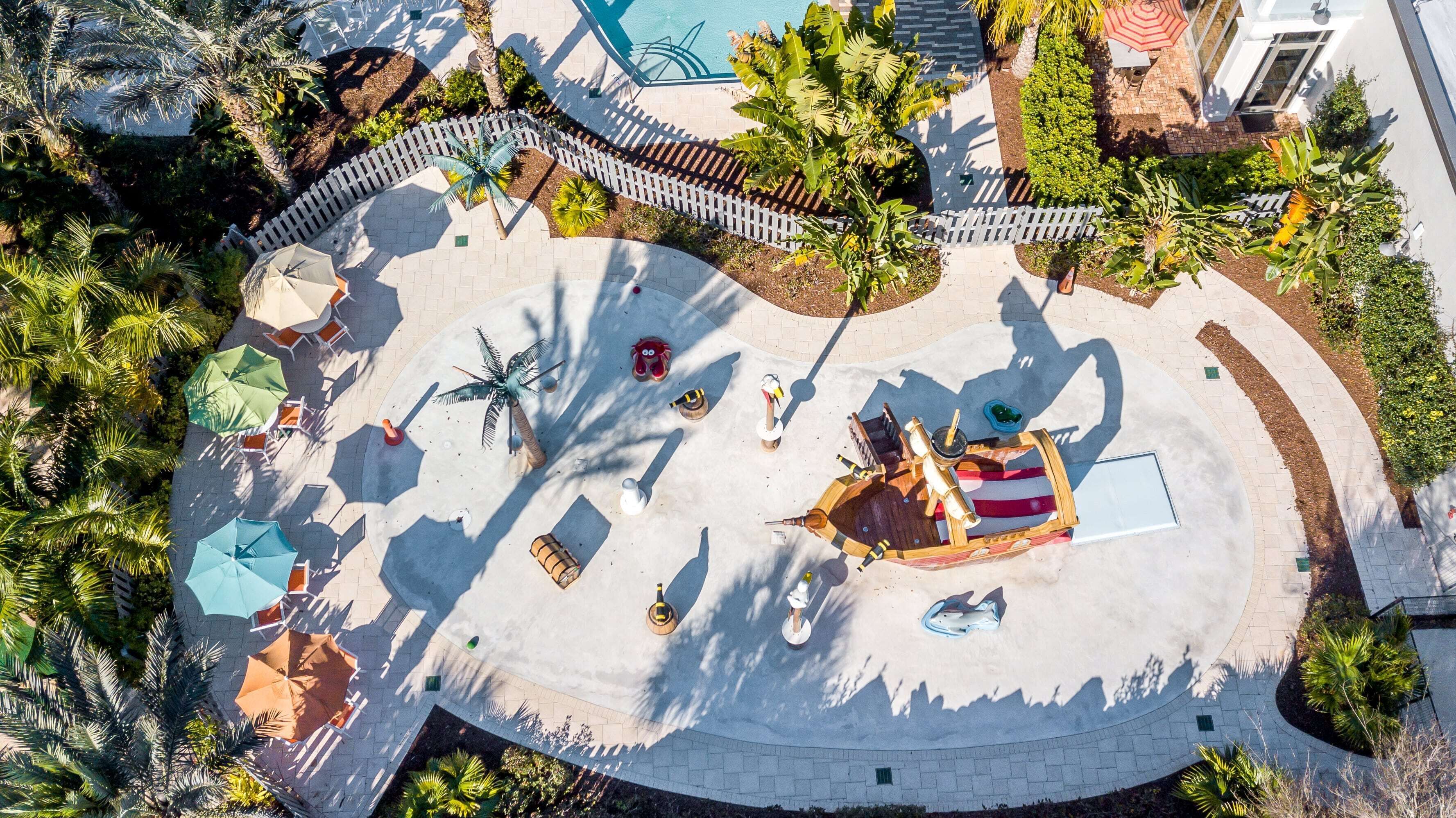 Vista aérea de una zona turística con piscina, palmeras y mobiliario exterior.