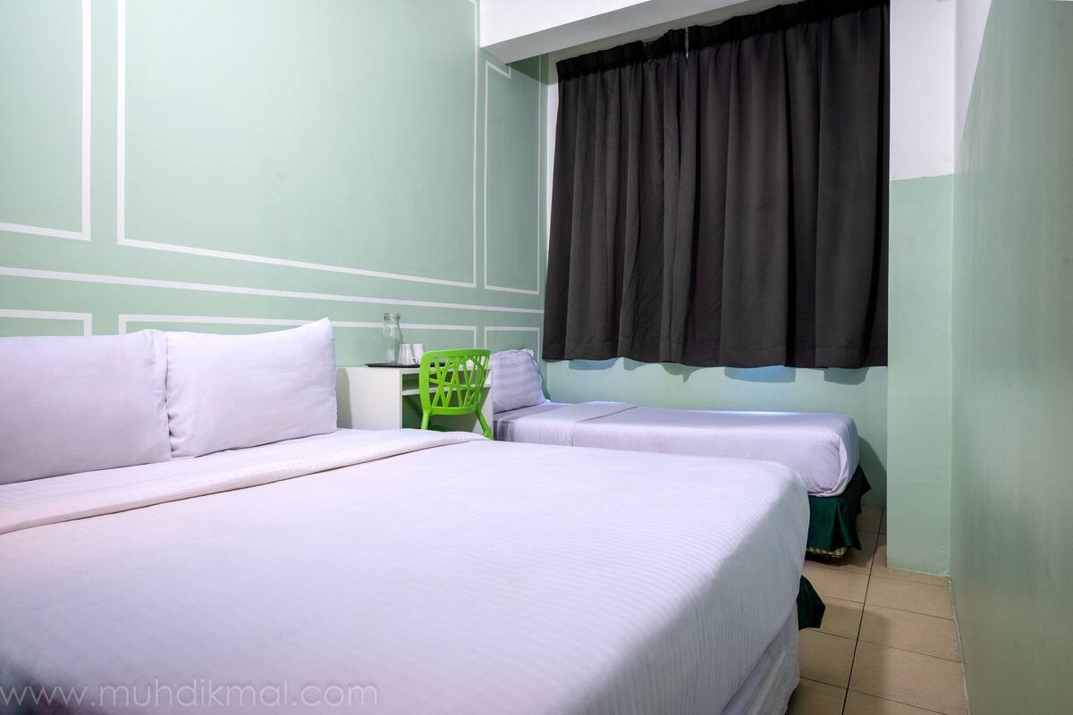 Een kamer met twee bedden, een groene stoel, een klein tafeltje met een fles en een raam met een zwart gordijn.
