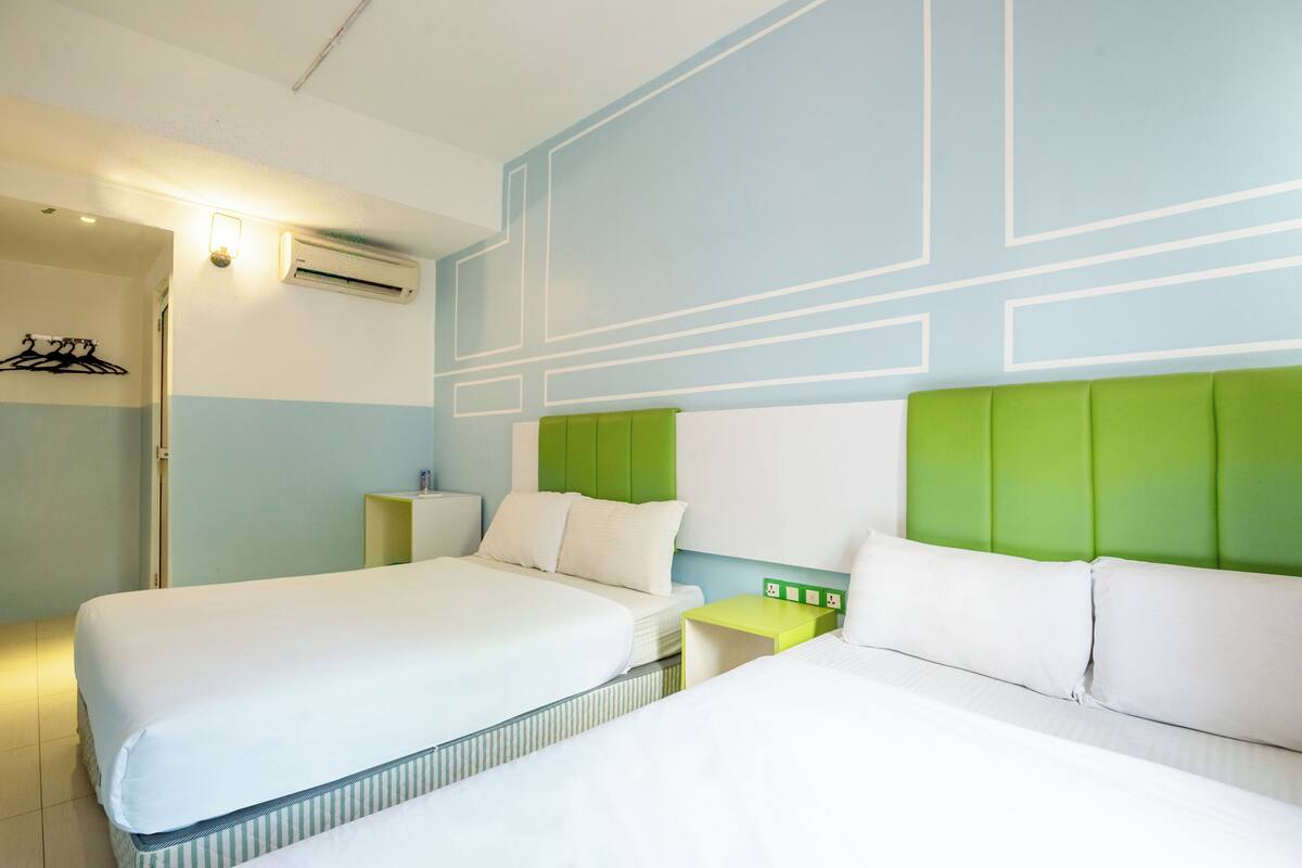 Een hotelkamer met twee bedden, een groen hoofdeinde, een nachtkastje en een wandairconditioner.