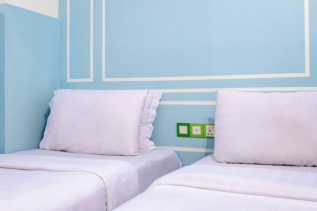 Een kamer met twee bedden, elk voorzien van witte beddengoed en kussens. Er zit een groene stopcontact in de muur.