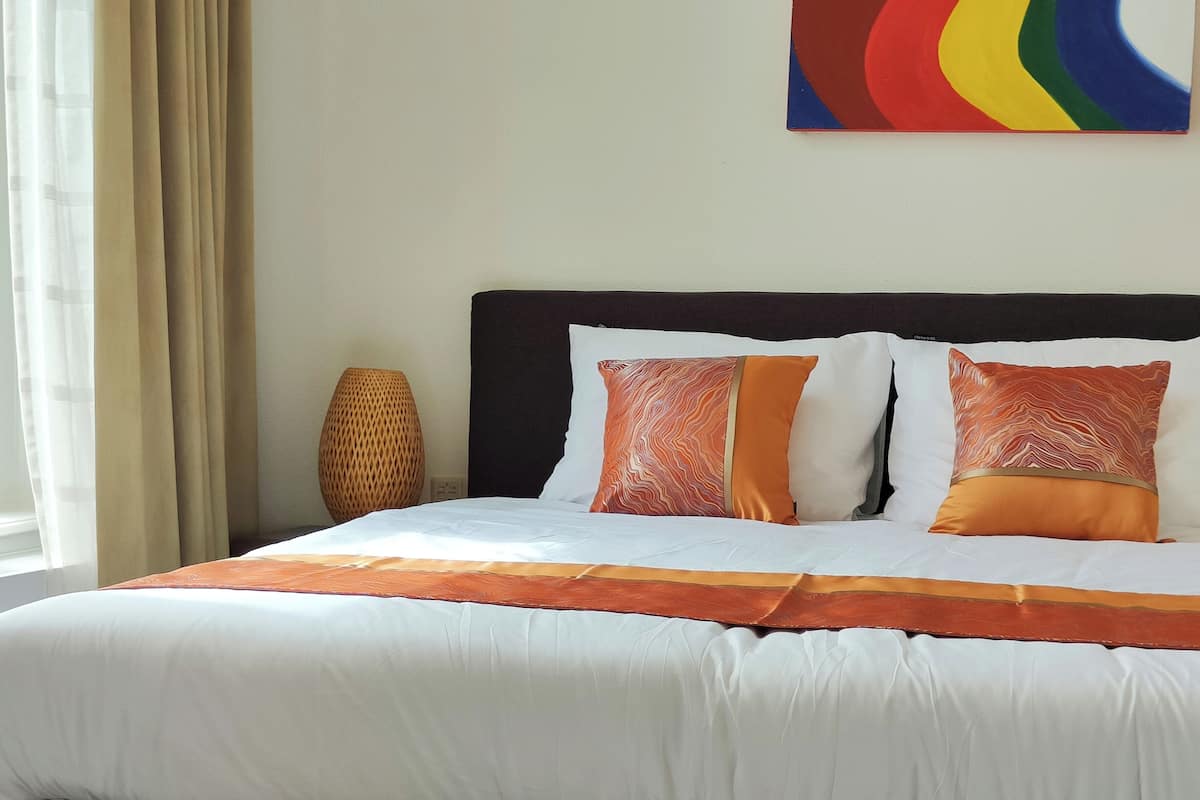 Un lit bien fait, avec une literie blanche et orange, une tête de lit sombre et, au-dessus, un tableau abstrait aux couleurs vives.