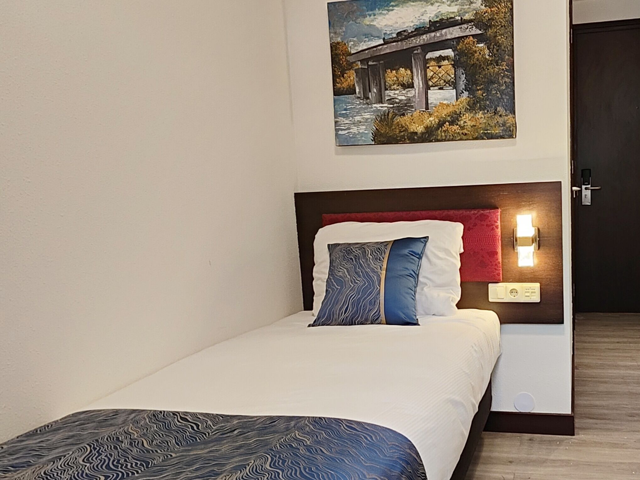 Una cama bien tendida con un edredón estampado, un almohadón con diseño ondulado y un cuadro de un puente en la pared.
