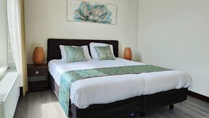Un dormitorio ordenado con una cama, dos mesitas de noche, una obra de arte en la pared y un ventanal con cortinas.