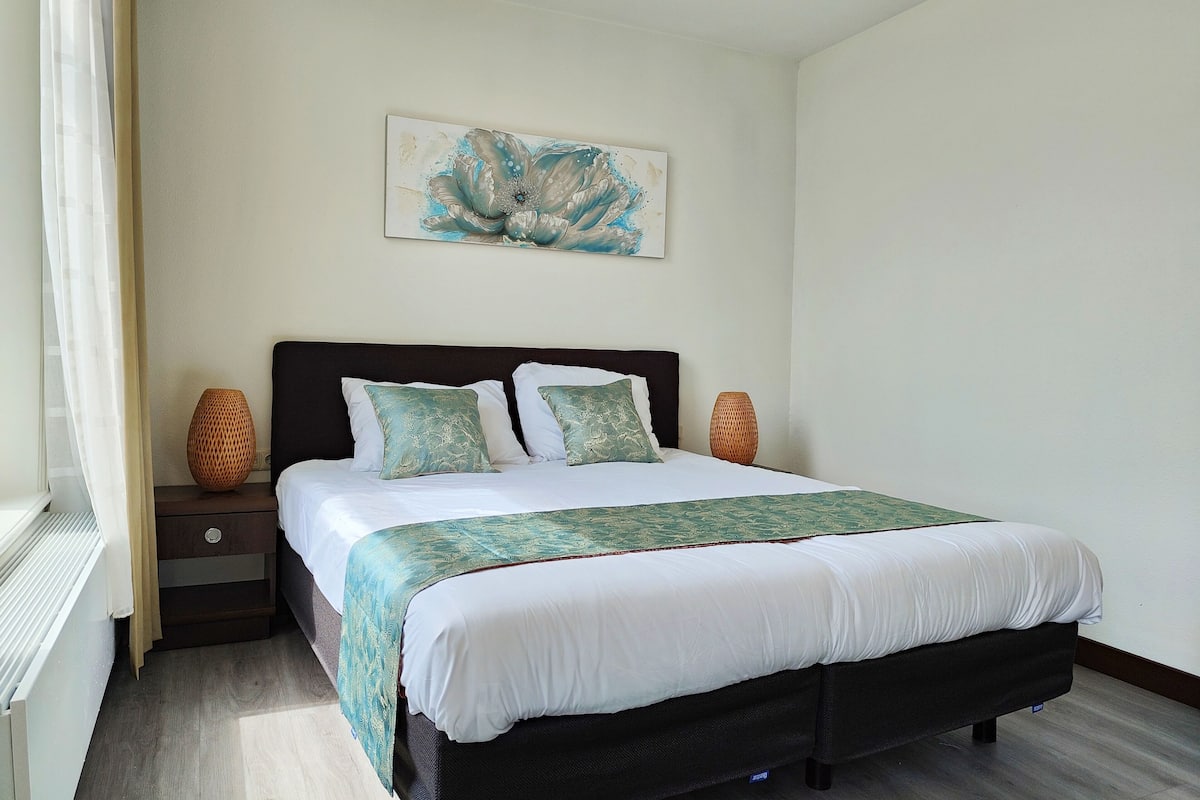 Une chambre à coucher bien rangée, avec un lit, deux tables de chevet, une œuvre d’art accrochée au mur et une fenêtre avec des rideaux.