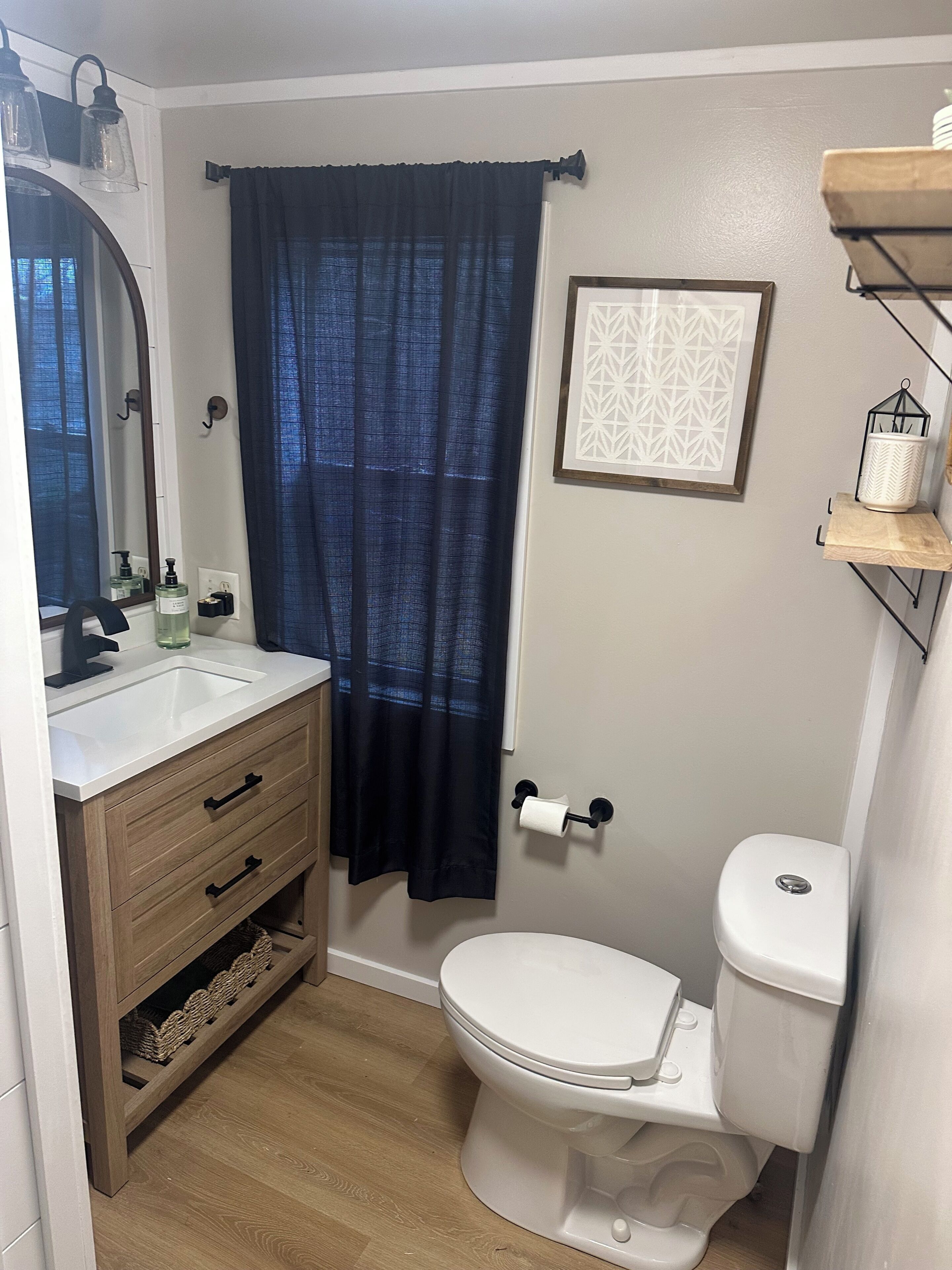 Ein Badezimmer mit weißem Waschbecken, Holz-Badmöbeln, einem dunkelblauen Duschvorhang, Toilette und einem rechteckigen Wandbild.