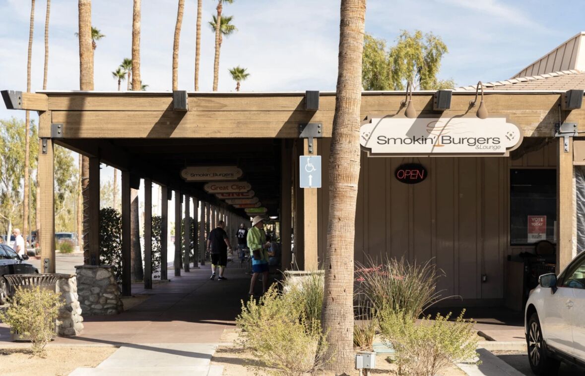 An outdoor seating area with a covered walkway, palm trees, and a sign for Smokin' Burgers & Lounge.