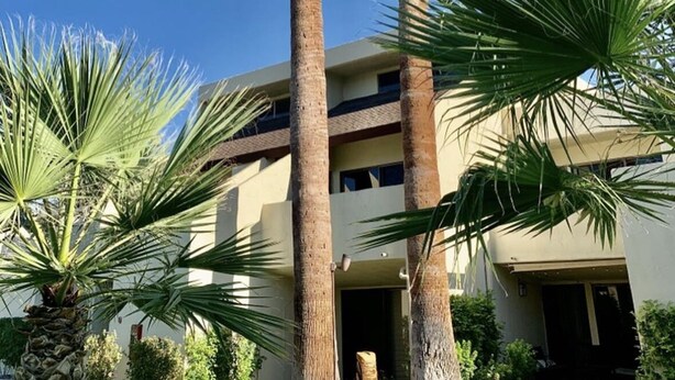 A building with two tall palm trees in front, a clear blue sky, and a beige facade. - Smoke Tree Racquet Club Condo 7 min Drive to Downtown Palm Springs!  (Palm Springs)