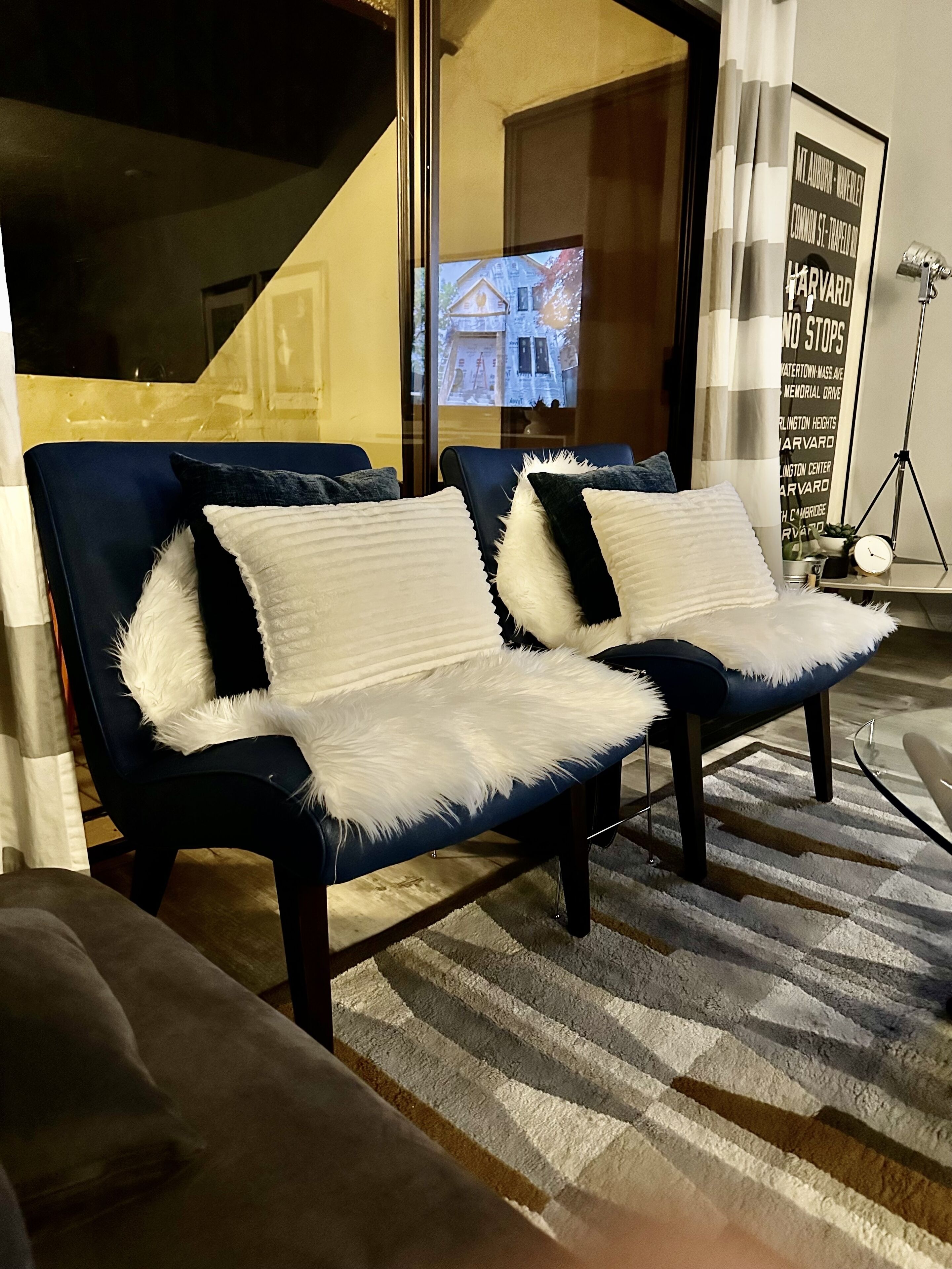 A modern living room with two dark blue sofas, each with a white fluffy pillow. A striped rug covers the floor, and a framed sign is visible on the wall.