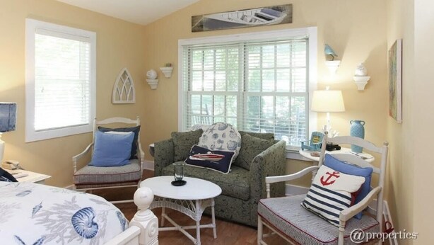 A cozy room with a green patterned sofa, a white armchair, and a small round table. There is a window with blinds, a lamp, and a decorative wall piece. - Charming New Buffalo Cottage (New Buffalo)